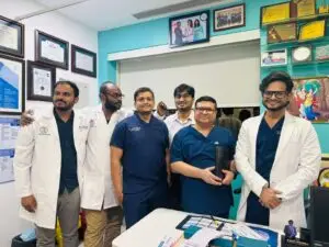 Six medical professionals, several in white coats and blue scrubs, pose together in a clinic with framed certificates on the wall behind them.