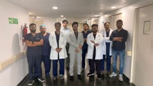 Group of doctors and medical staff posing in a hospital corridor, some in scrubs and white coats with arms crossed or relaxed poses.
