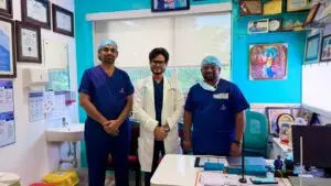 Three healthcare workers stand in a clinic, two in blue scrubs and a doctor in a white coat, smiling in front of a wall lined with certificates and a desk cluttered with supplies.